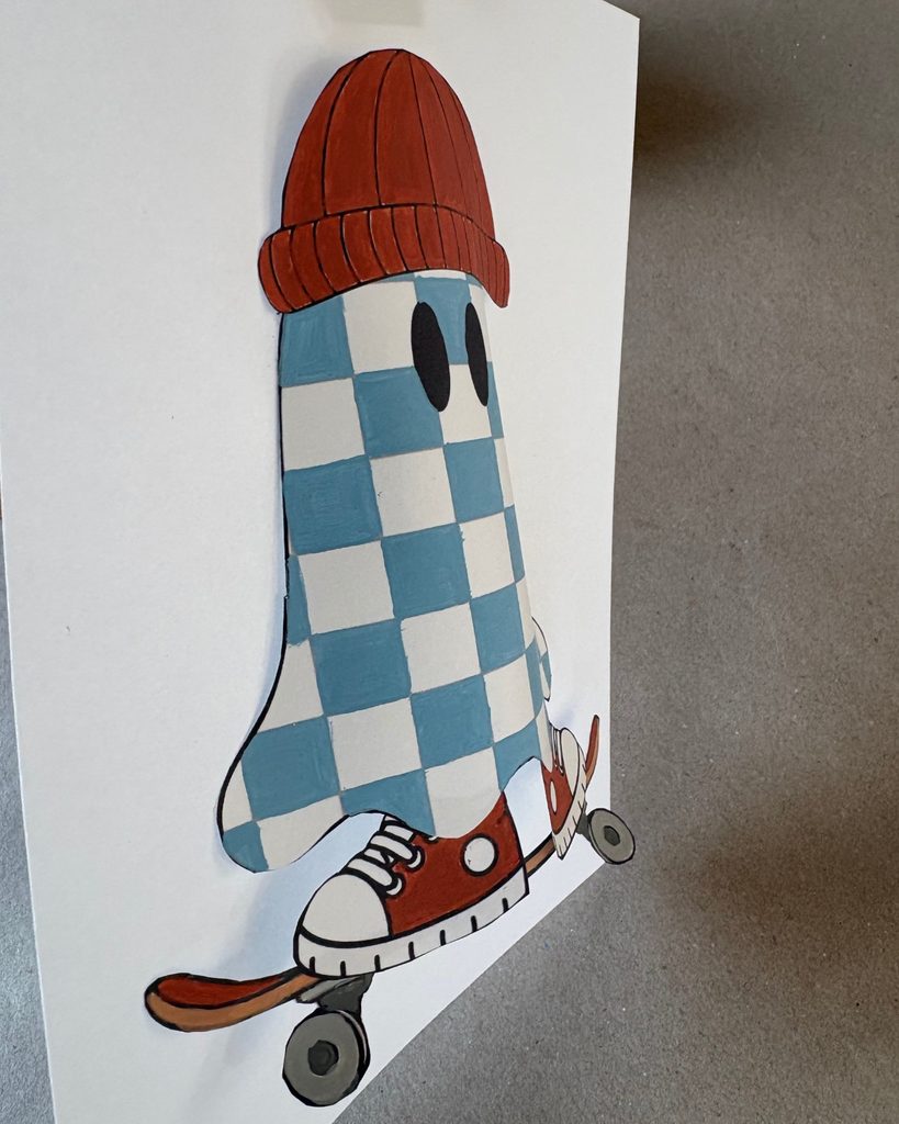 Side view of a blue checkered 3D paper ghost craft wearing a red beanie and sneakers on a skateboard, showing its curved pop-out shape.
