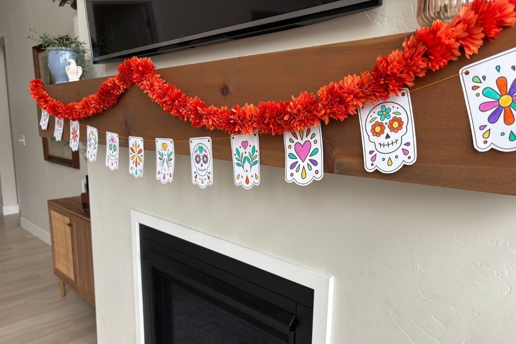 Colorful DIY Día de los Muertos paper banner with skulls, hearts, and flowers hanging across a fireplace mantel.