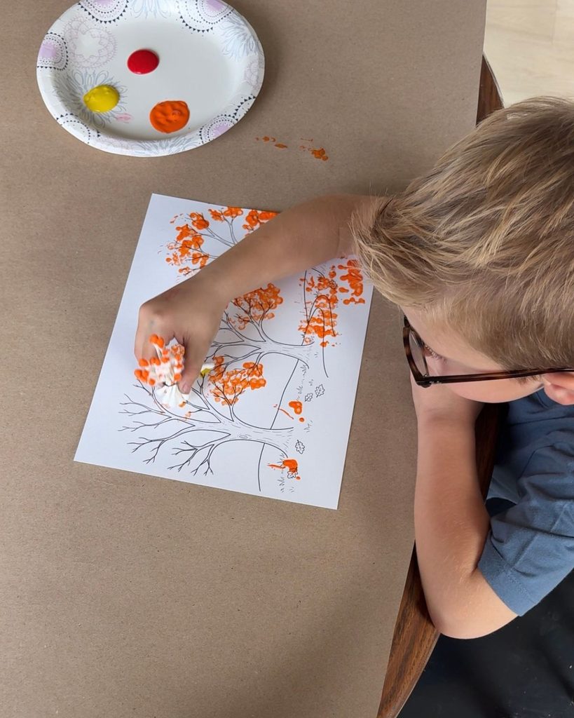 Child using a bundle of Q-tips to paint orange fall leaves on a printable tree.